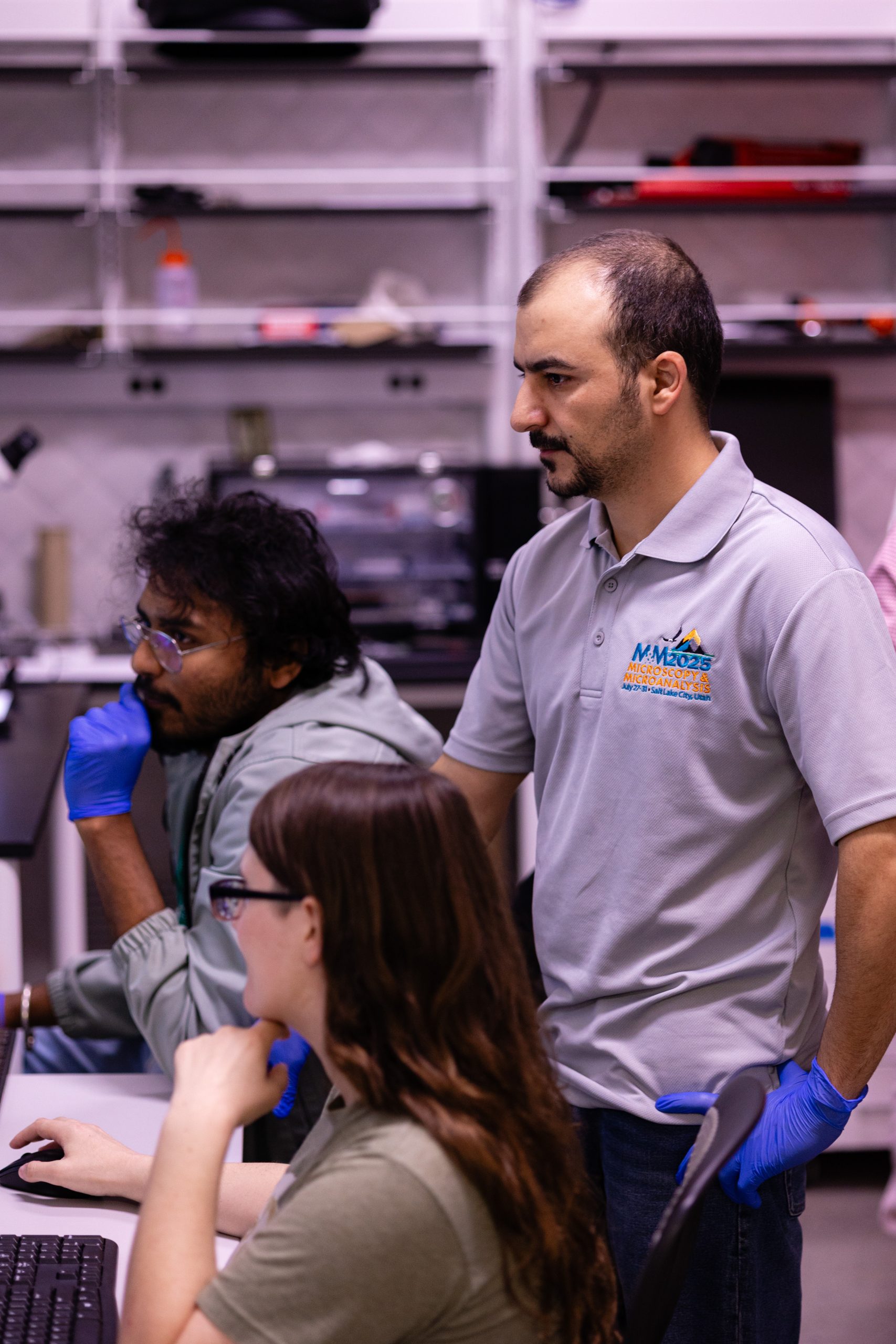 A man in a "Microscopy and Microanalysis 2025" shirt observes students' work