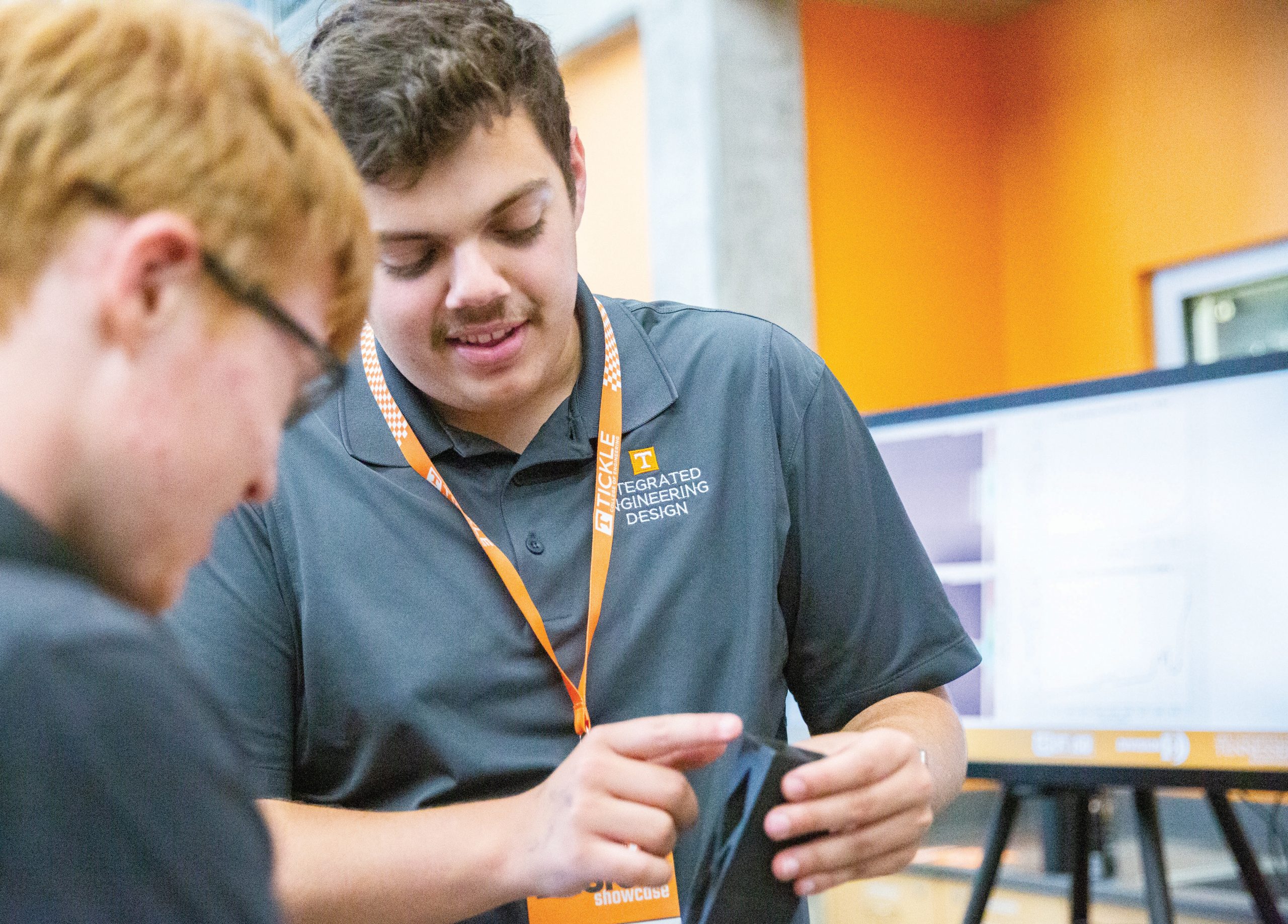 A senior design team member shows a mechanism piece to a visitor