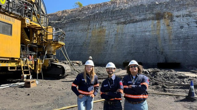 Three UT students pose in hard hats in front of a high-wall miner